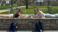 Two students on a stonewall on campus with laptops.