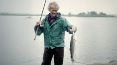 Dort Cameron stands in a body of water wearing fishing gear and holding a fish.