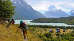 Backpackers hike along a trail with a river and mountains in the background.