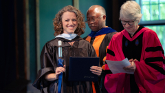 A Bread Loaf School of English student wearing academic regalia smiles and holds her Middlebury cane.