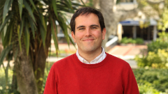 A professor wearing a red sweater poses for a photo outside.