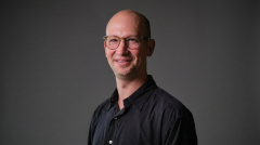 A professor in a black shirt and glasses smiles at the camera.