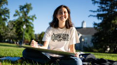 A student wearing a white t-shirt and sitting outside smiles at the camera while working on a drawing.