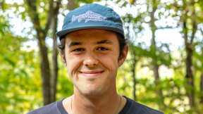 A student with a blue baseball hat smiles at the camera while standing in the woods.