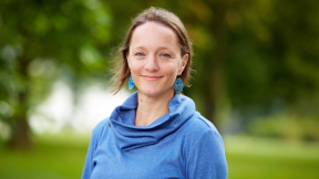 Outdoor photo portrait of Mez Baker-Medard wearing a blue shirt with green grass and trees behind. 