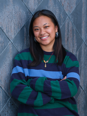 A student in a dark sweater leans up against a patterned wall outside.