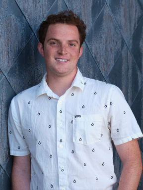A student in a white shortsleeved shirt leans against a patterned wall outside.