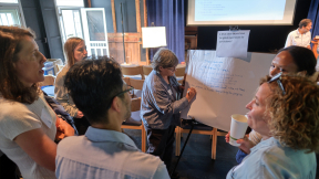 A group of people brainstorm around a flip chart with one person taking notes.