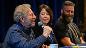 A person speaks into a microphone at a table during a panel discussion.