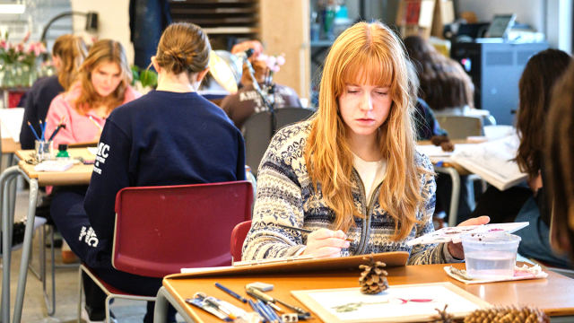 A student draws in a sketchbook at a classroom table. 
