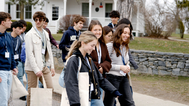 A group of students wearing jackets and carrying tote bags walks across a college campus.