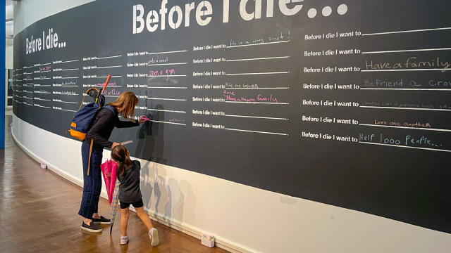 A parent and child stand at a large blackboard in a hallway.