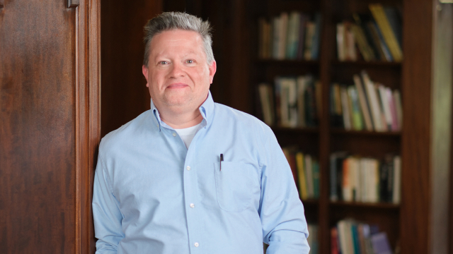 Photo of an adult in a light blue shirt leaning against a wall with books in the background.