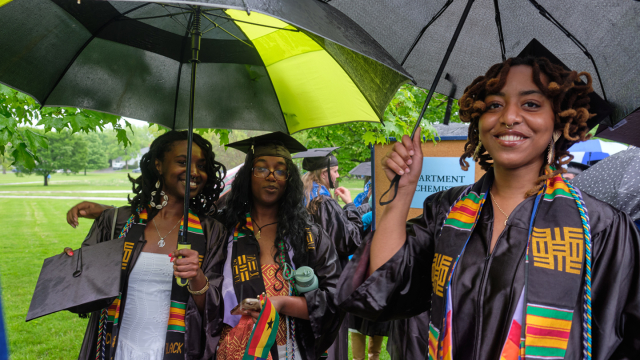 Students stand and smile under umbrellas at Commencement.