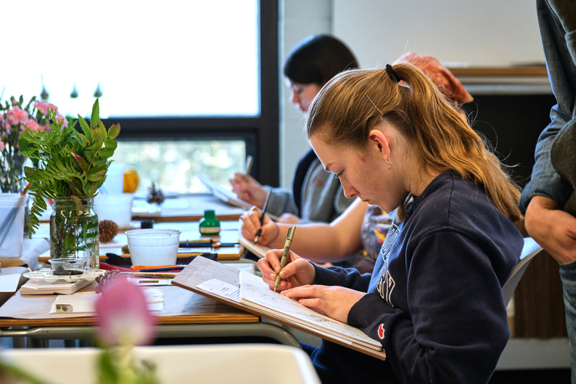 A student sketches in a book at a classroom table. 