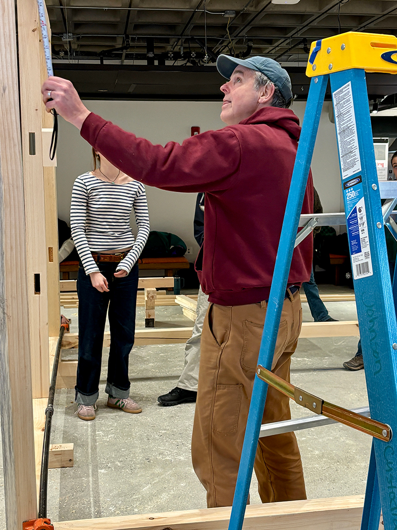 Man in red sweatshirt measures a wall of a timber structure.