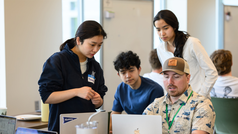 A professor wearing a shortsleeve shirt and ball cap speaks with students at laptop computers.
