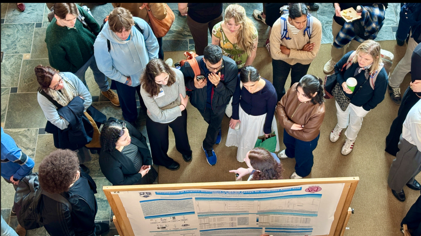 A group of students listen to a presenter at a poster session.