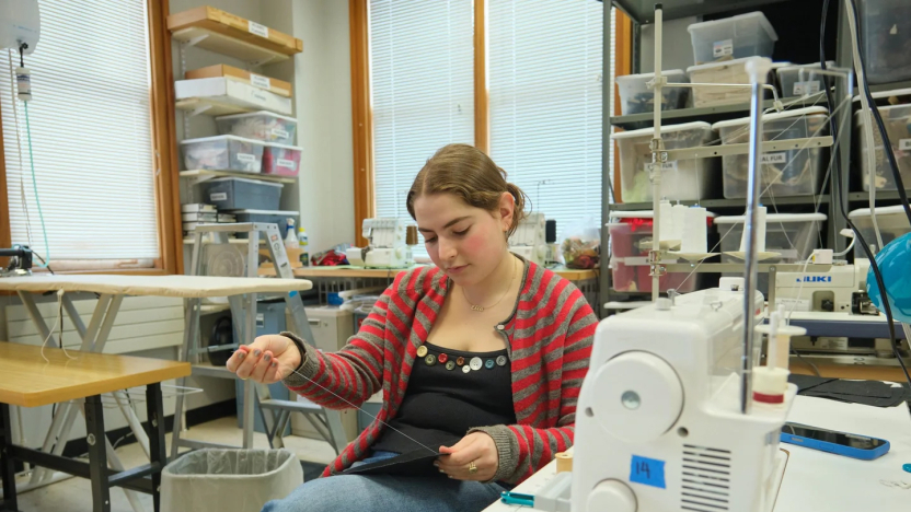 A student sits and sews by hand surrounded by costume materials.