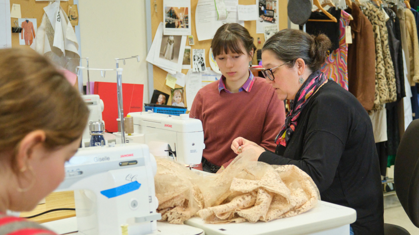 Two people sit at a sewing machine in a costume shop.