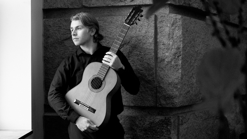 A student stands against a stone wall inside holding a guitar and looking out a window.