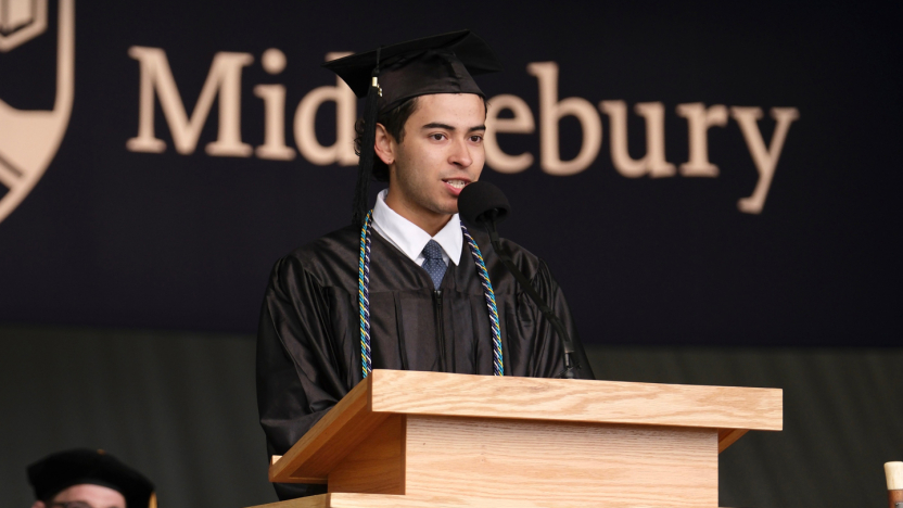 A student in cap and gown gives a speech at a podium with a Middlebury banner in the background..