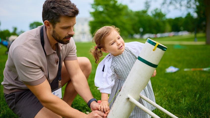 An adult and young child load a t-shirt cannon on a grassy lawn.