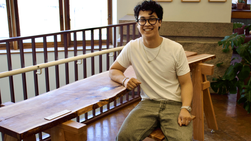 A student wearing a white tee shirt poses for a photo while sitting at a table.