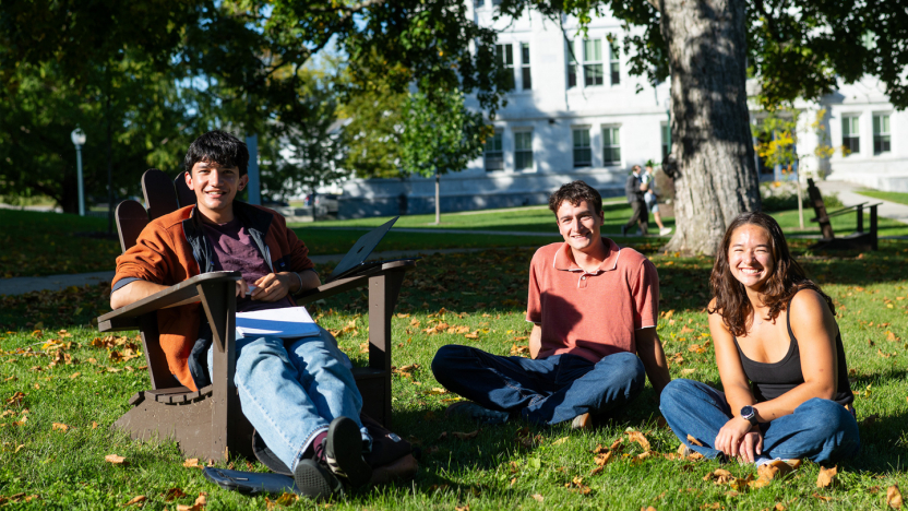Three students sit on the lawn in front of a campus building, one in an Adirondack chair.