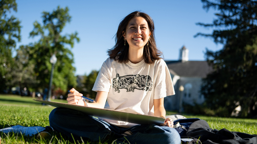 A student wearing a white t-shirt and sitting outside smiles at the camera while working on a drawing.