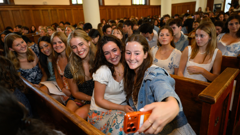 Students in chapel pews take a group selfie surrounded by smiling classmates.