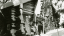 A historic black and white photo of the Hubbard log cabin from the 1950s with two students with skis in front of the door.