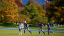 A woman in a Middlebury jersey hits a field hockey ball with a stick. Three opposing players are trailing closely behind her. 