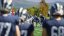The foreground of the photo is blurry, looking through a group of football players on the sidelines. The background is in focus, and you see a formation of football players on the field ready to snap into action. 